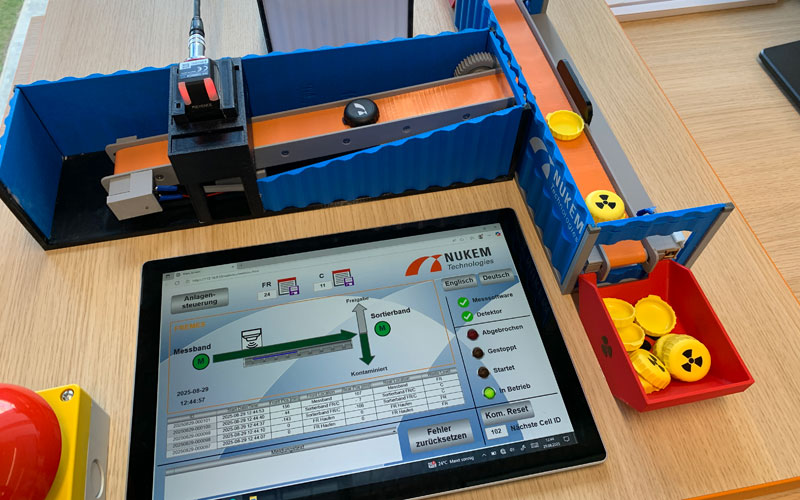 Ein Modell einer Sortieranlage mit Förderband und Bildschirm zeigt die Trennung radioaktiv markierter Objekte anhand eines Steuerungssystems.